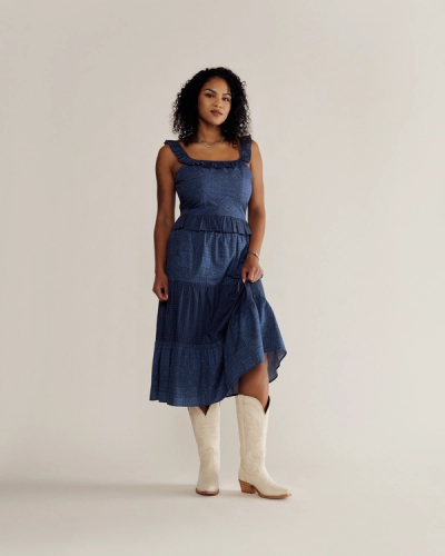 A woman in a blue sleeveless dress and white cowboy boots stands against a plain beige background, looking at the camera.