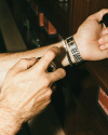 A person sprays perfume on their wrist while wearing a metal watch, a black beaded bracelet, and another bracelet with text. In the background are wooden shelves with books.