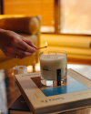 A hand lighting a candle with a match. The candle is placed on top of a book titled "Same Pa." The background shows a room with warm lighting.