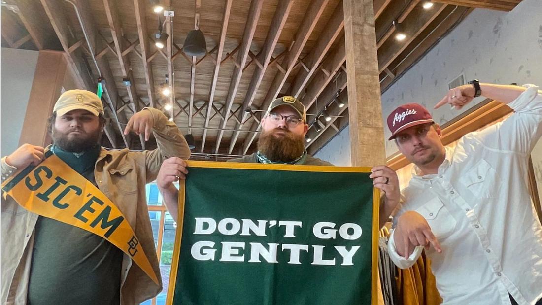Three men stand indoors; the man on the left holds a "Sic 'em" banner, the middle man holds a "Don't Go Gently" sign, and the man on the right points downward, each displaying different hats.