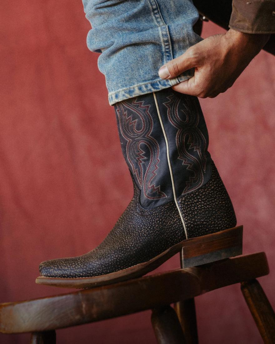 A person wearing blue jeans and a dark, patterned cowboy boot rests their foot on a wooden chair against a reddish background.