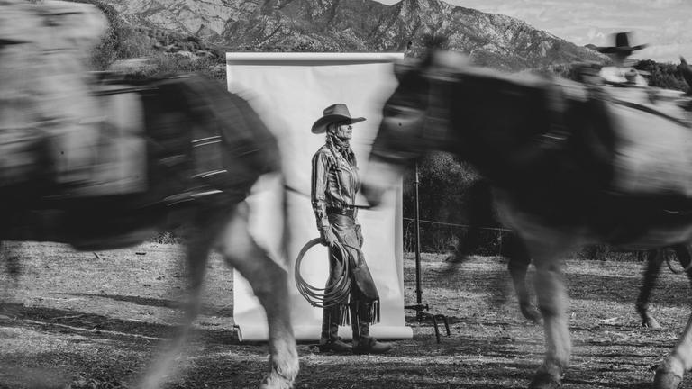 A person in cowboy attire stands in front of a white backdrop outdoors, holding a lasso, while two blurred horses pass by; mountains are visible in the background.
