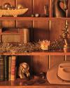 A wooden bookshelf displays a cowboy hat, books, a vintage radio, candles, a horse bust, deer head, boots, a bowl, coasters, and metallic garland.