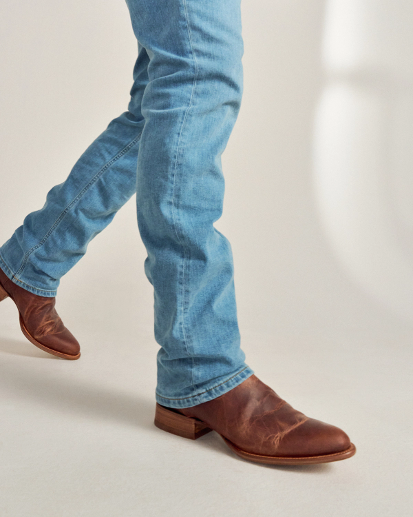 Person wearing light blue jeans and brown leather boots walking on a light-colored surface.