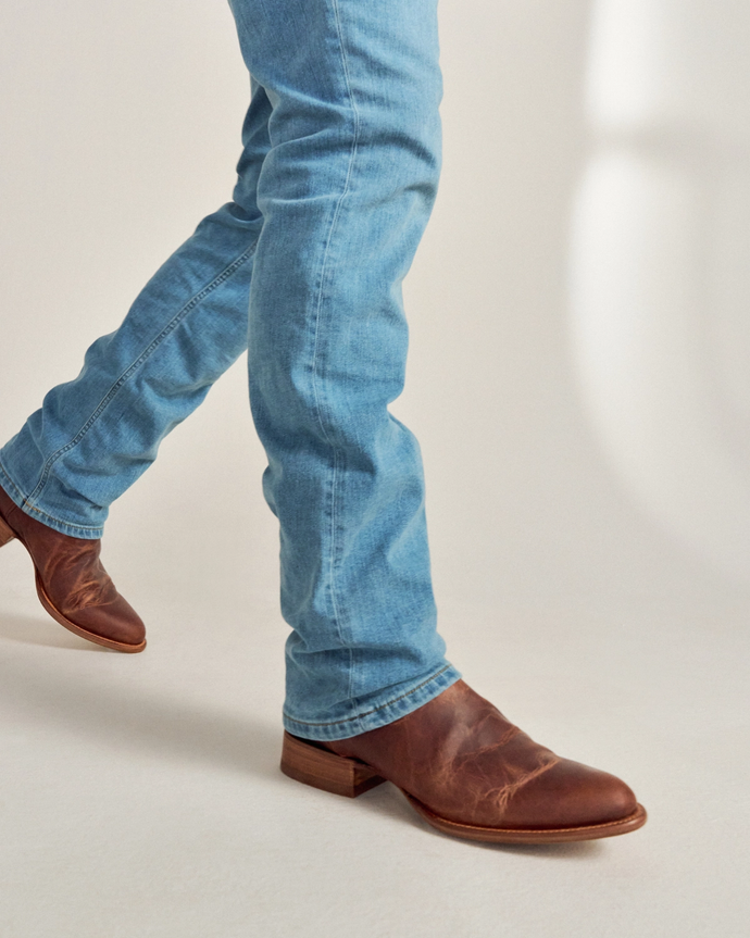 Person wearing light blue jeans and brown leather boots walking on a light-colored surface.