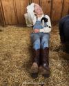 @WOMANGONEWEST Woman wearing brown Annie boots sits in hay in a barn while holding a baby goat in her overalls.
