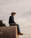 A man wearing a cowboy hat, denim shirt, and jeans sits on a large, weathered metal structure outdoors, looking into the distance.