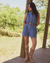A woman stands on a wooden deck wearing a denim romper, a red belt, and brown cowboy boots, with trees and greenery in the background.