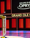 A microphone stands on a branded pedestal on stage at the Grand Ole Opry, with the venue's name illuminated in the background.