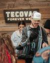 A man wearing a Tecovas hat and striped jacket inspects a boot at a store counter, with other people and a Tecovas sign in the background.
