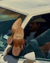 A person wearing cowboy boots with “TECVNAS” and “TECVAS” on the soles rests their feet out the window of a car; the person wears a cowboy hat and sits in the driver’s seat.