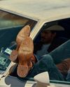 A person wearing cowboy boots with “TECVNAS” and “TECVAS” on the soles rests their feet out the window of a car; the person wears a cowboy hat and sits in the driver’s seat.
