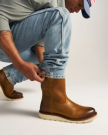 The Knox in Wheat Cowhide. A person wearing light blue jeans and brown wheat-colored cowhide leather boots squats down, adjusting one pant leg. Only the lower half of the body is visible.