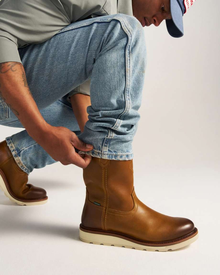 The Knox in Wheat Cowhide. A person wearing light blue jeans and brown wheat-colored cowhide leather boots squats down, adjusting one pant leg. Only the lower half of the body is visible.
