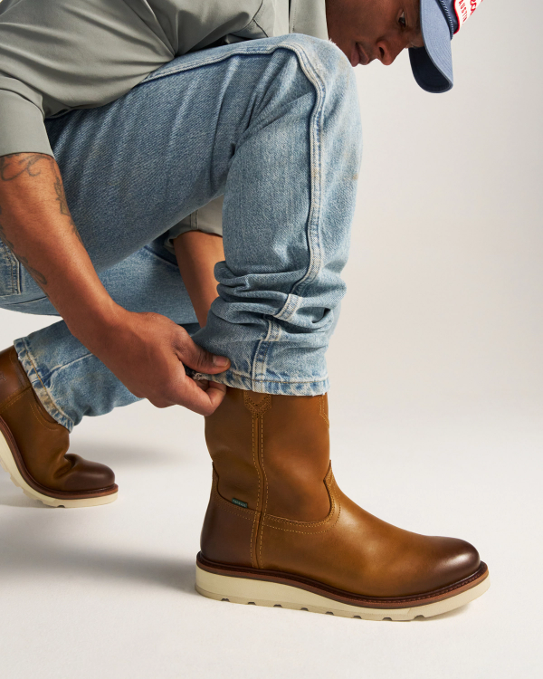 The Knox in Wheat Cowhide. A person wearing light blue jeans and brown wheat-colored cowhide leather boots squats down, adjusting one pant leg. Only the lower half of the body is visible.