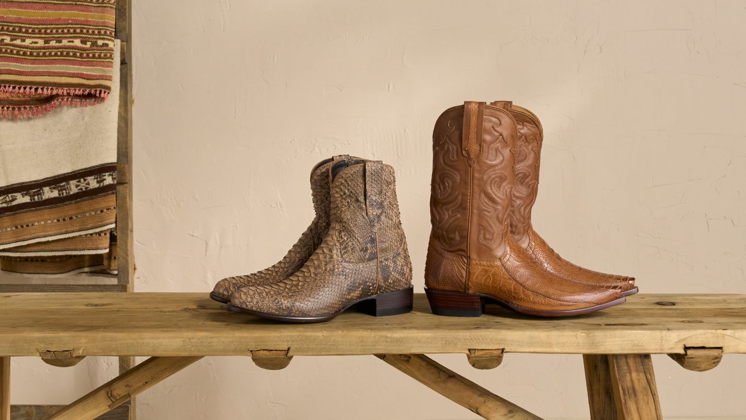 Two pairs of cowboy boots on a wooden table. One pair is brown with intricate patterns, and the other pair has a textured, snakeskin-like appearance. A woven textile hangs in the background.