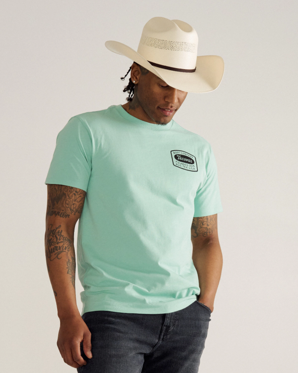 A person wearing a light blue T-shirt, dark jeans, and a white cowboy hat stands with one hand in their pocket, looking downward against a plain background.