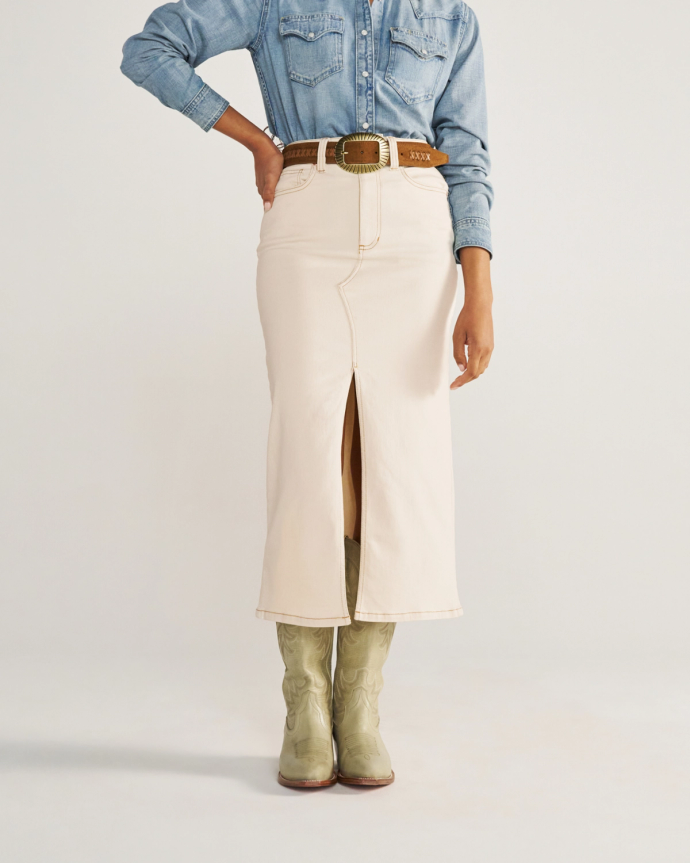 Person wearing a light denim shirt, cream-colored skirt with a front slit, tan belt with a large buckle, and light green cowboy boots.