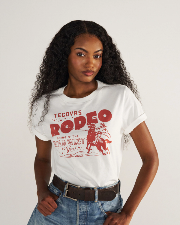 A woman with long curly hair wears a white T-shirt with a red "Tecovas Rodeo" graphic, blue jeans, and a brown belt, standing against a plain background.