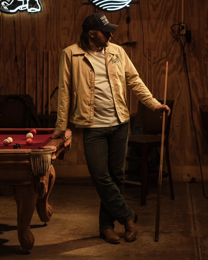 Man in a beige jacket and jeans holding a pool cue, leaning against a pool table with balls scattered, in a dimly lit room.