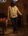 Man in a beige jacket and jeans holding a pool cue, leaning against a pool table with balls scattered, in a dimly lit room.