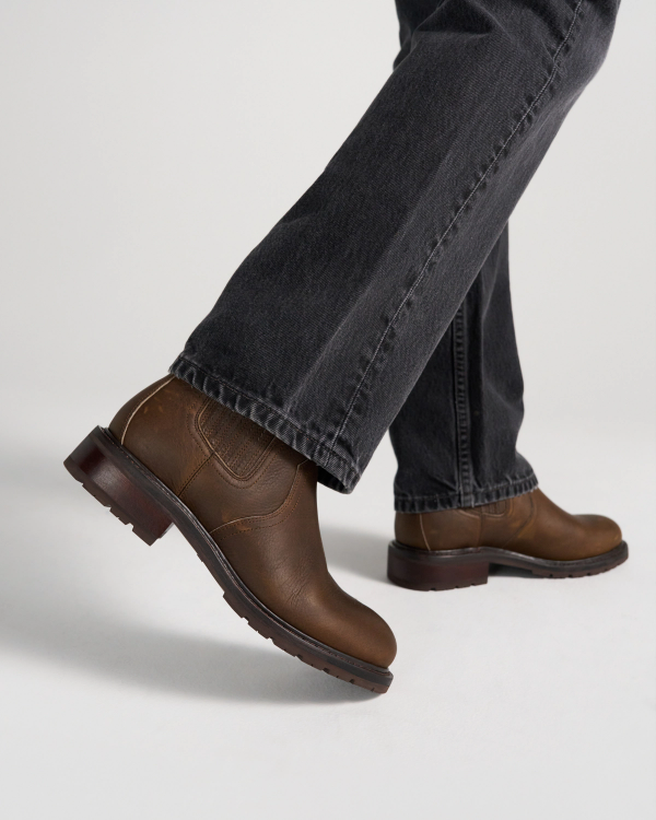 The Chelsea in Cafe Cowhide. A person wearing dark jeans and brown leather boots with cafe cowhide accents is walking on a white surface.