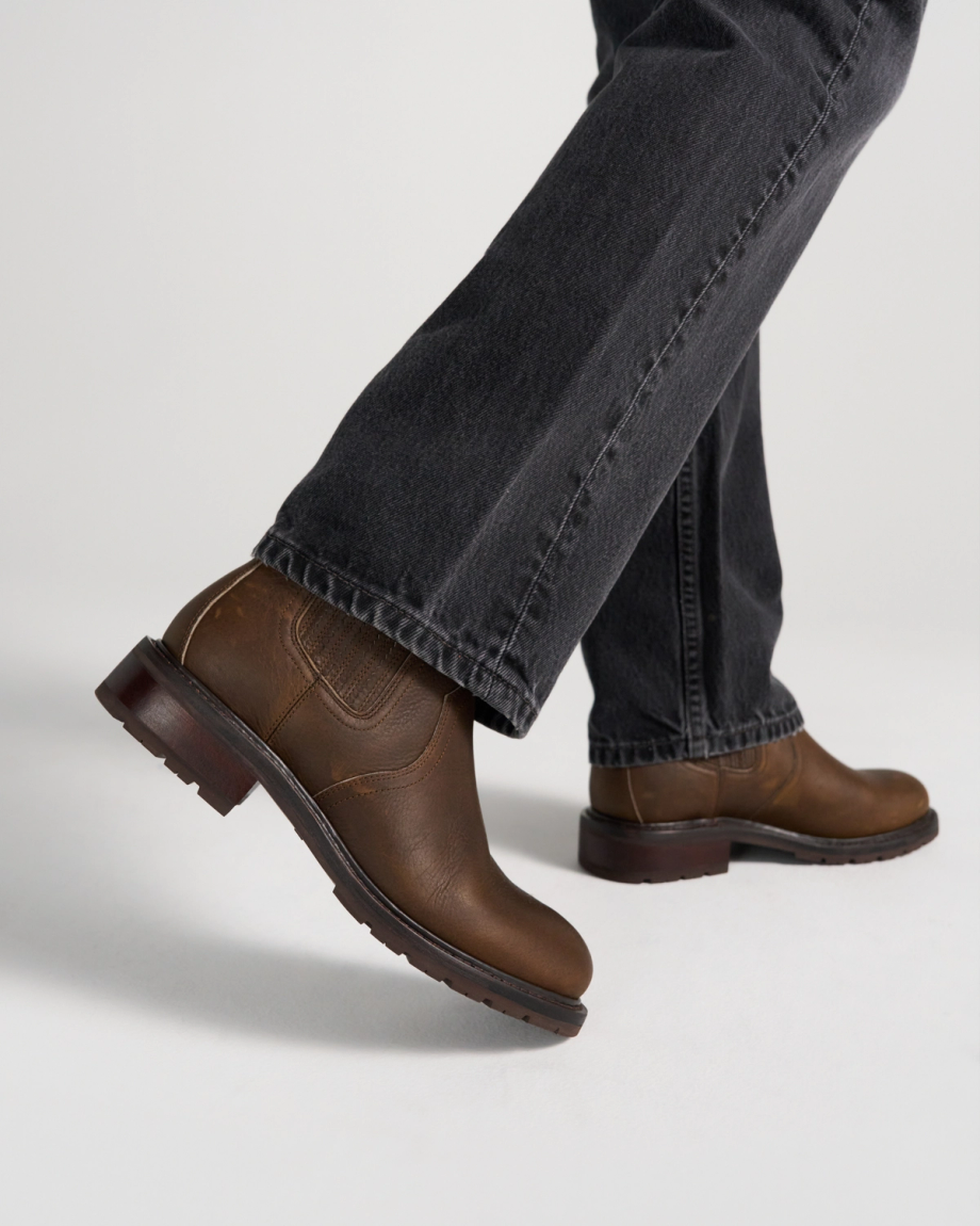 The Chelsea in Cafe Cowhide. A person wearing dark jeans and brown leather boots with cafe cowhide accents is walking on a white surface.