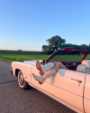 A person wearing white cowboy boots relaxes with their legs hanging out of the passenger side of a white convertible car on a rural road at sunset.