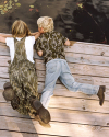Two children lie on a wooden dock, looking into the water. One wears camouflage overalls and boots; the other wears a camouflage shirt, jeans, and brown shoes. Lily pads float nearby.