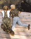 Two children lie on a wooden dock, looking into the water. One wears camouflage overalls and boots; the other wears a camouflage shirt, jeans, and brown shoes. Lily pads float nearby.