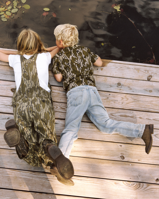 Two children lie on a wooden dock, looking into the water. One wears camouflage overalls and boots; the other wears a camouflage shirt, jeans, and brown shoes. Lily pads float nearby.