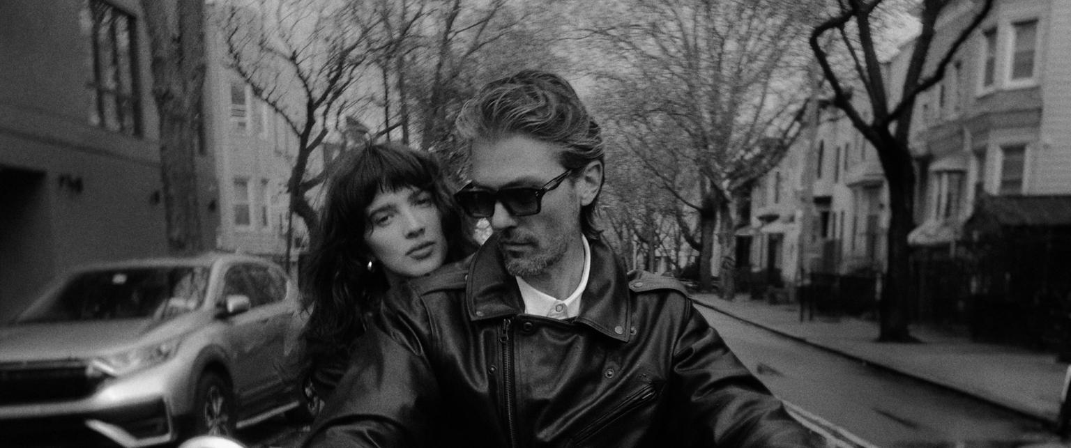 A woman and man wearing leather jackets ride a motorcycle down a city street lined with trees and parked cars.