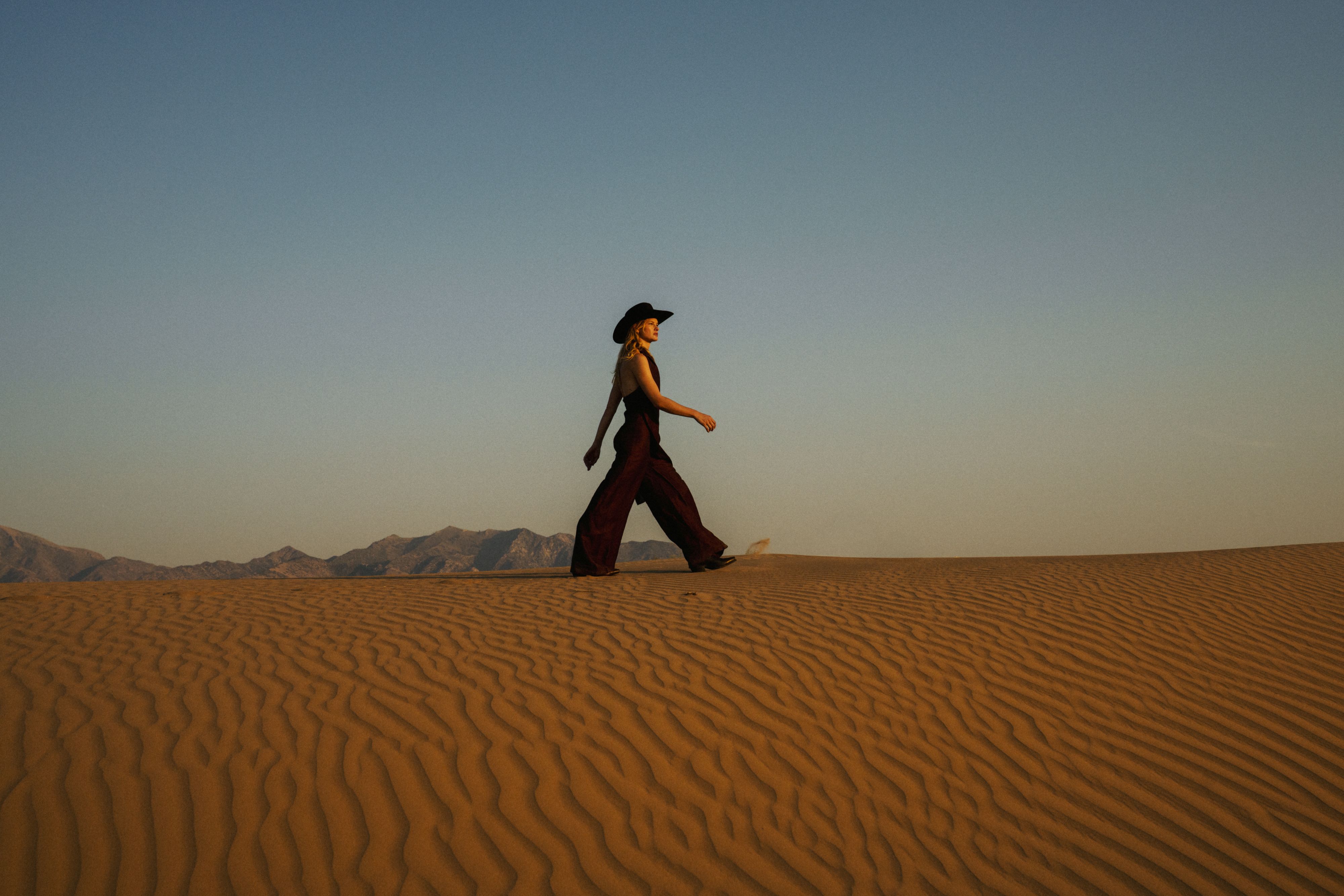 A woman in a wide-brimmed hat and dark jumpsuit walking across a sand dune at sunset.