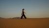 A woman in a wide-brimmed hat and dark jumpsuit walking across a sand dune at sunset.