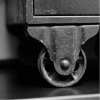 Close-up black and white image of a metal caster wheel and corner bracket attached to a textured object, possibly a cart or vintage furniture.