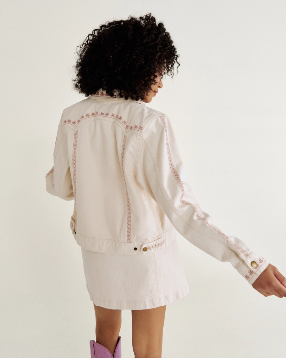 Person with curly hair wearing a cream jacket with pink embroidery and a matching skirt, stands against a plain background, holding the jacket open slightly Natural Denim .