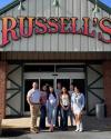 Seven people stand in front of the entrance to a building with a large sign reading "RUSSELL'S" above them.