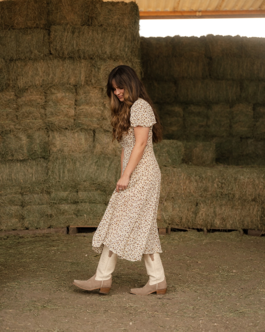 A woman in a floral dress and boots walks in front of stacked hay bales in a barn.