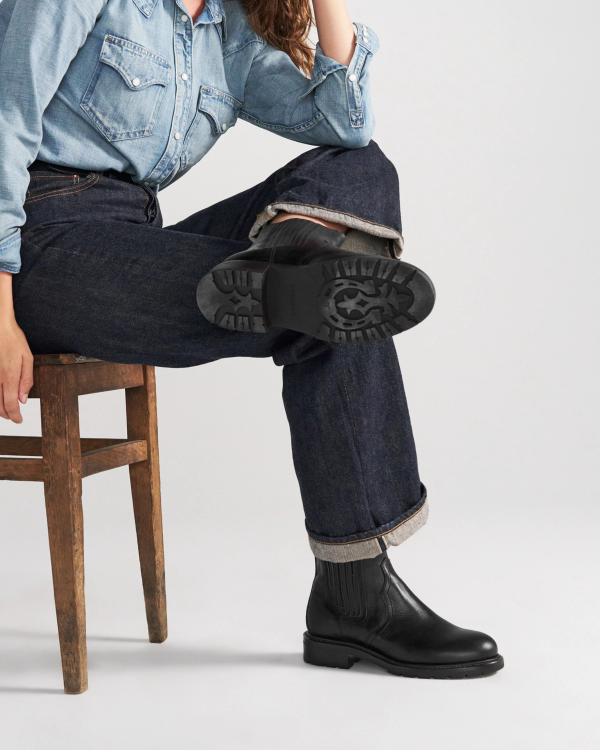 The Chelsea in Midnight Cowhide. A person in a denim shirt and jeans sits on a wooden chair with one leg crossed, showing the sole of their black Midnight Cowhide boot.