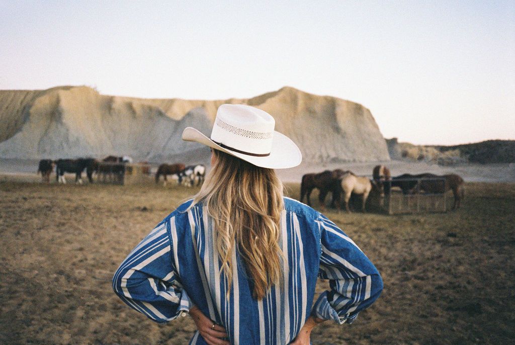 A person in a cowboy hat and striped shirt stands with hands on hips, facing a herd of horses near a rocky landscape.