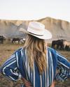 A person in a cowboy hat and striped shirt stands with hands on hips, facing a herd of horses near a rocky landscape.