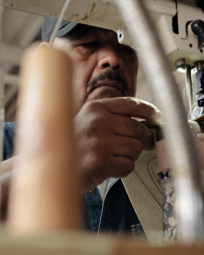 A man wearing a cap operates a sewing machine, focusing on threading or adjusting the fabric under the needle.