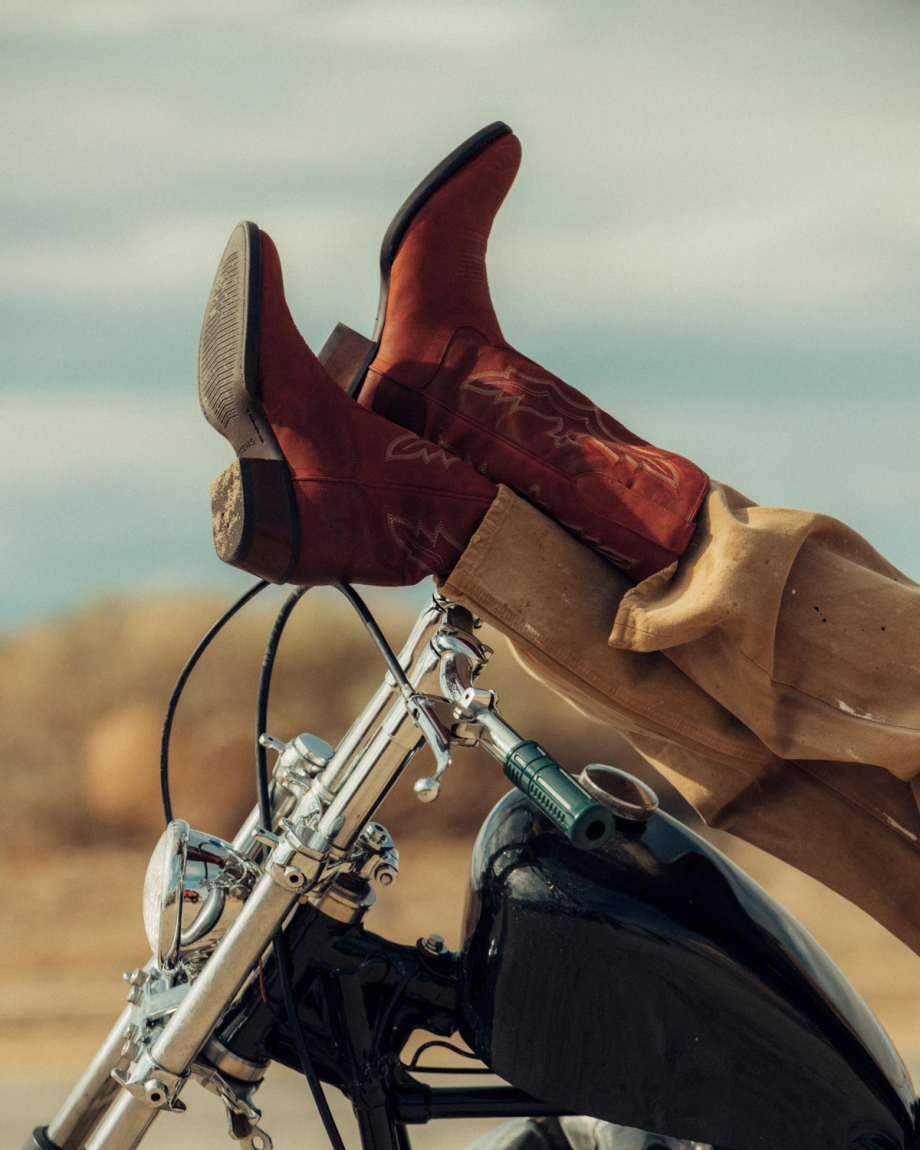 The Britt in Redwood Goat. Person wearing brown cowboy boots and tan pants rests their feet on the handlebars of a black motorcycle, surrounded by redwood trees in an outdoor setting.