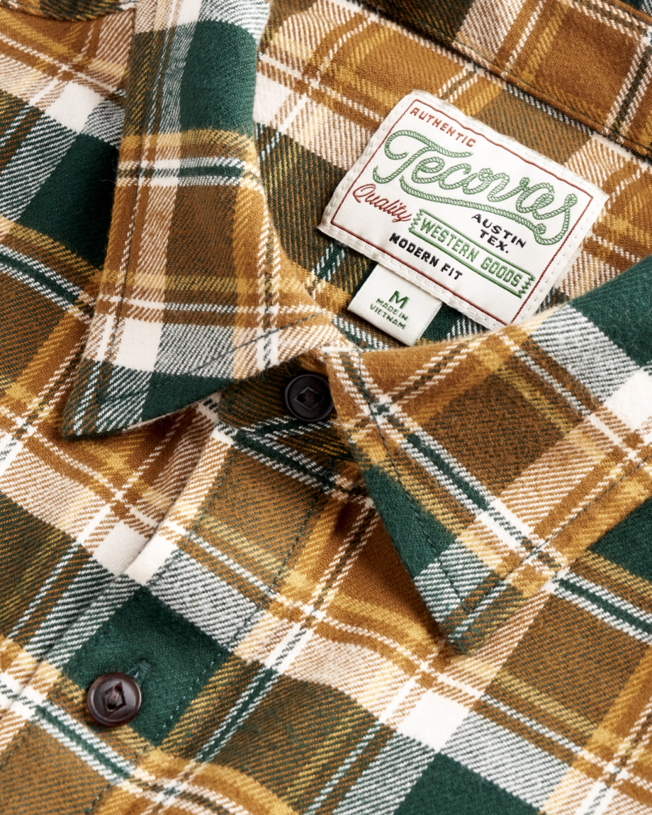 Close-up of a green, brown, and white plaid flannel shirt with dark buttons and a sewn label reading “Jeans Authentic Quality Western Goods” and “Modern Fit.”.