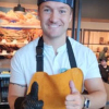 Person wearing a blue cap, yellow apron, and black glove, giving a thumbs up indoors.