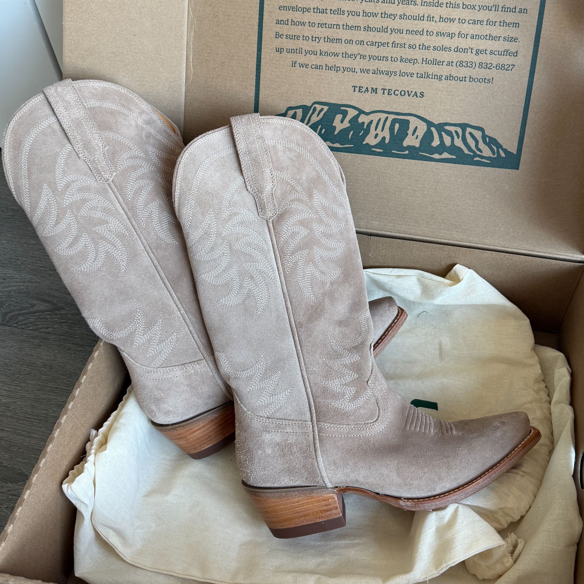 A pair of light beige suede cowboy boots with wooden heels displayed inside an open cardboard box.