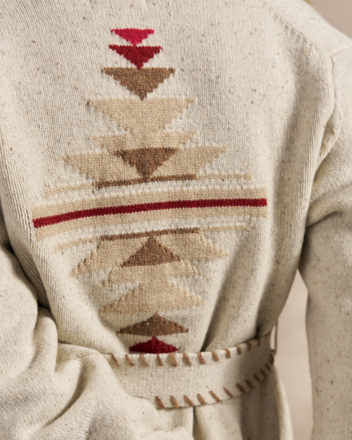 Back view of a person wearing a cream-colored knit sweater with a geometric, tribal-inspired pattern in shades of red and brown, and a matching belt.