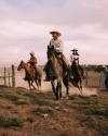 Three cowboys riding horses in a dusty field, with focused expressions, under a cloudy sky.
