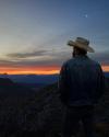 A man in a cowboy hat and denim jacket watches a sunset over a mountainous landscape with a crescent moon above.