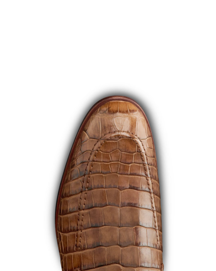 The Bennett in Saddle Alligator. Close-up of the toe of a brown leather shoe with a crocodile pattern, reminiscent of The Bennett Alligator, photographed against a plain white background.